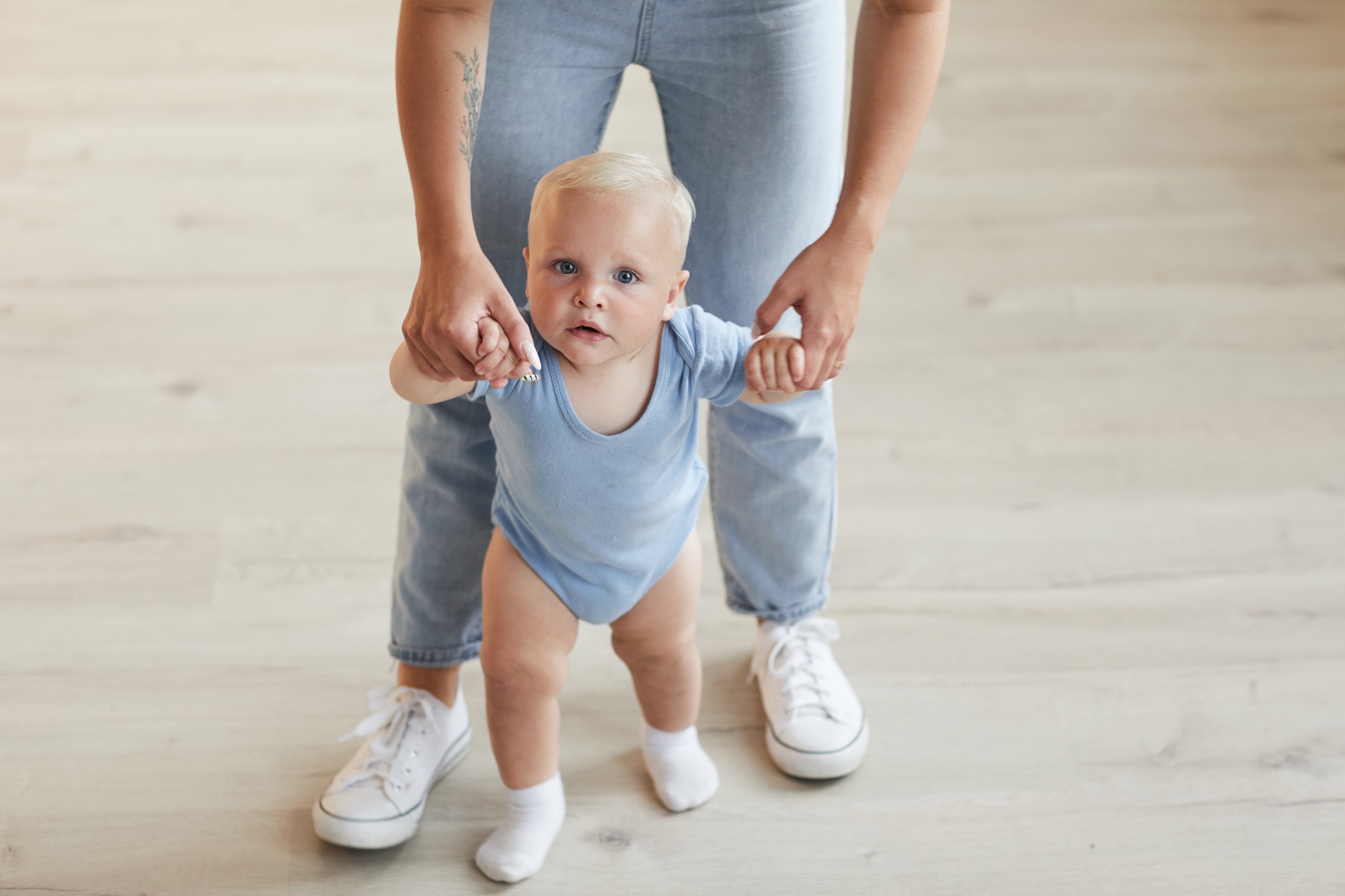 fisioterapia pediatrica Un bebé de pelo rubio, con un mono azul claro y calcetines blancos, es sostenido por las manos de un adulto desde atrás. El adulto lleva vaqueros azul claro y zapatillas blancas y está de pie sobre un suelo de madera clara. Daganzo Adyre