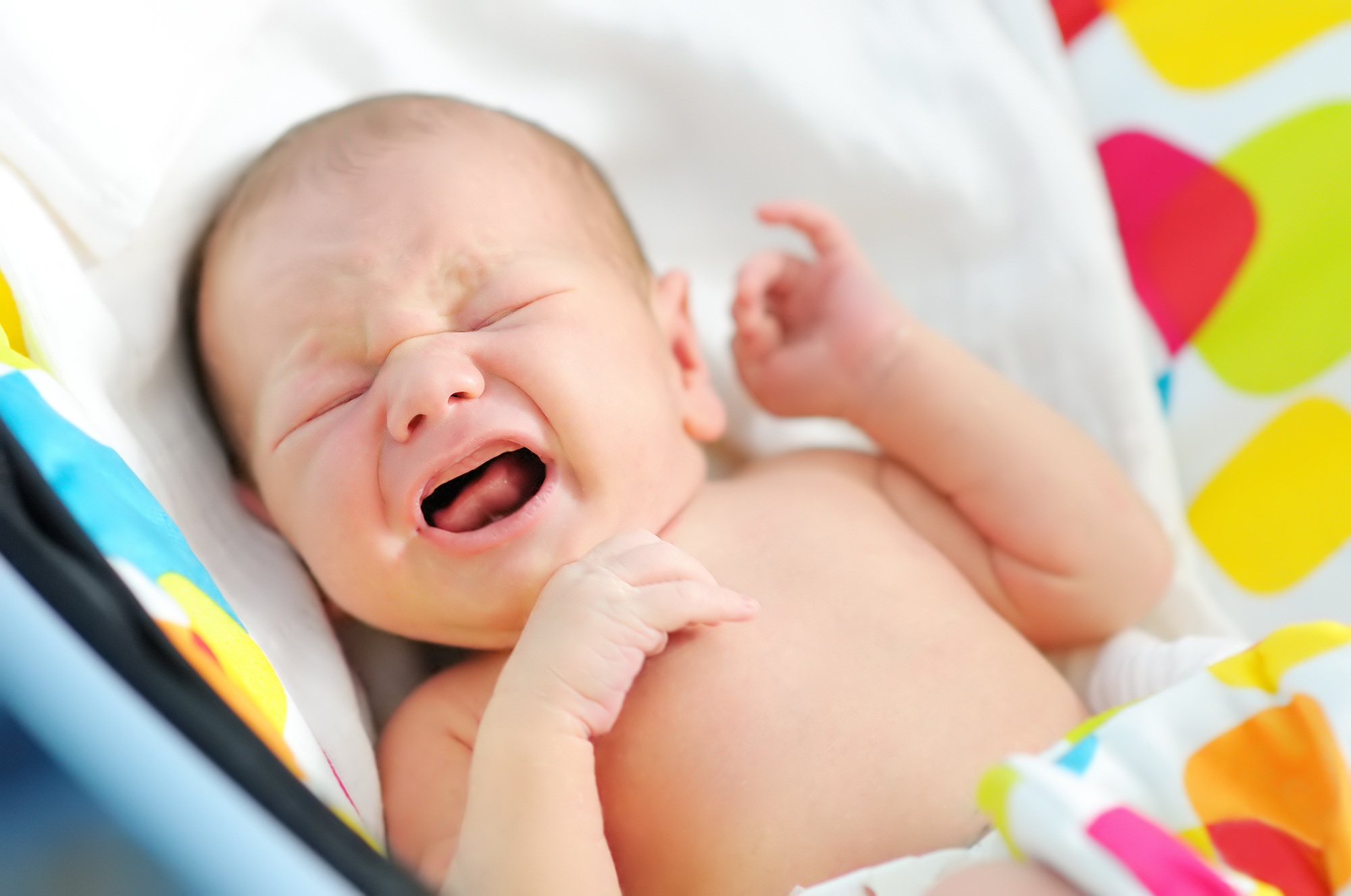 fisioterapia pediatrica Un bebé recién nacido llora, acostado sobre una manta con estampados de colores. Tiene los ojos bien cerrados y una mano levantada cerca de su cara. El fondo presenta colores brillantes que crean un ambiente vibrante. Daganzo Adyre