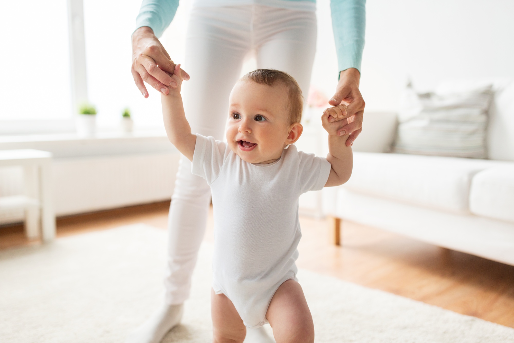 fisioterapia pediatrica Un bebé con un mono blanco camina alegremente de la mano de un adulto. La habitación está iluminada por la luz del sol y se ve un sofá al fondo. El bebé parece feliz y concentrado en dar sus pasos. Daganzo Adyre