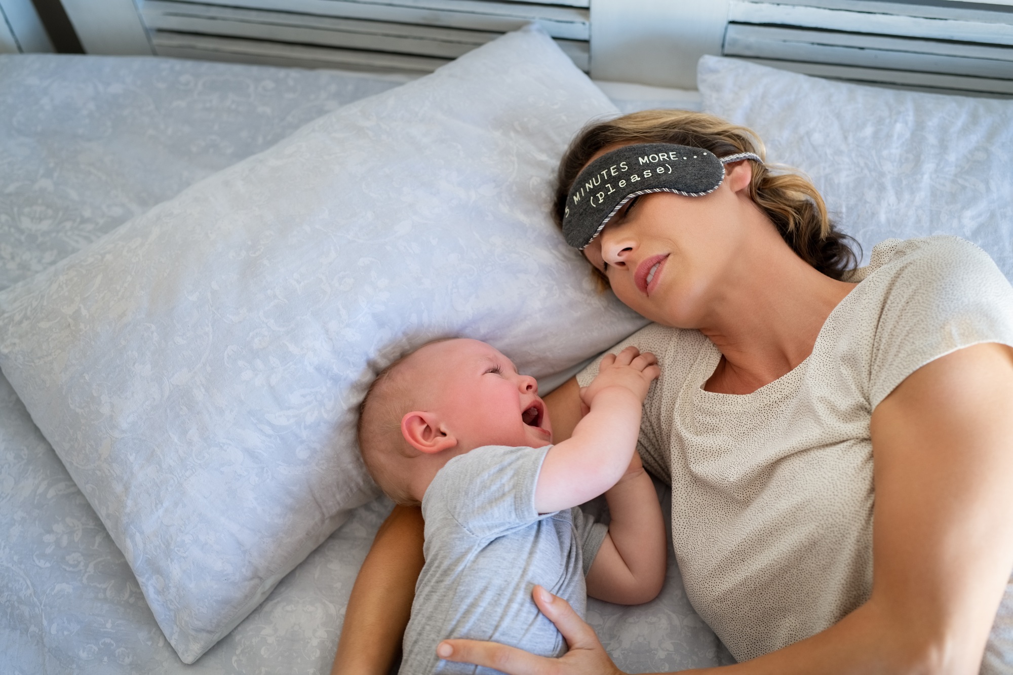 fisioterapia pediatrica Una mujer con un antifaz que dice "5 minutos más (por favor y gracias)" yace en una cama junto a un bebé que se ríe y trata de alcanzarla. La escena sugiere un momento relajado y lúdico entre ellos. Daganzo Adyre