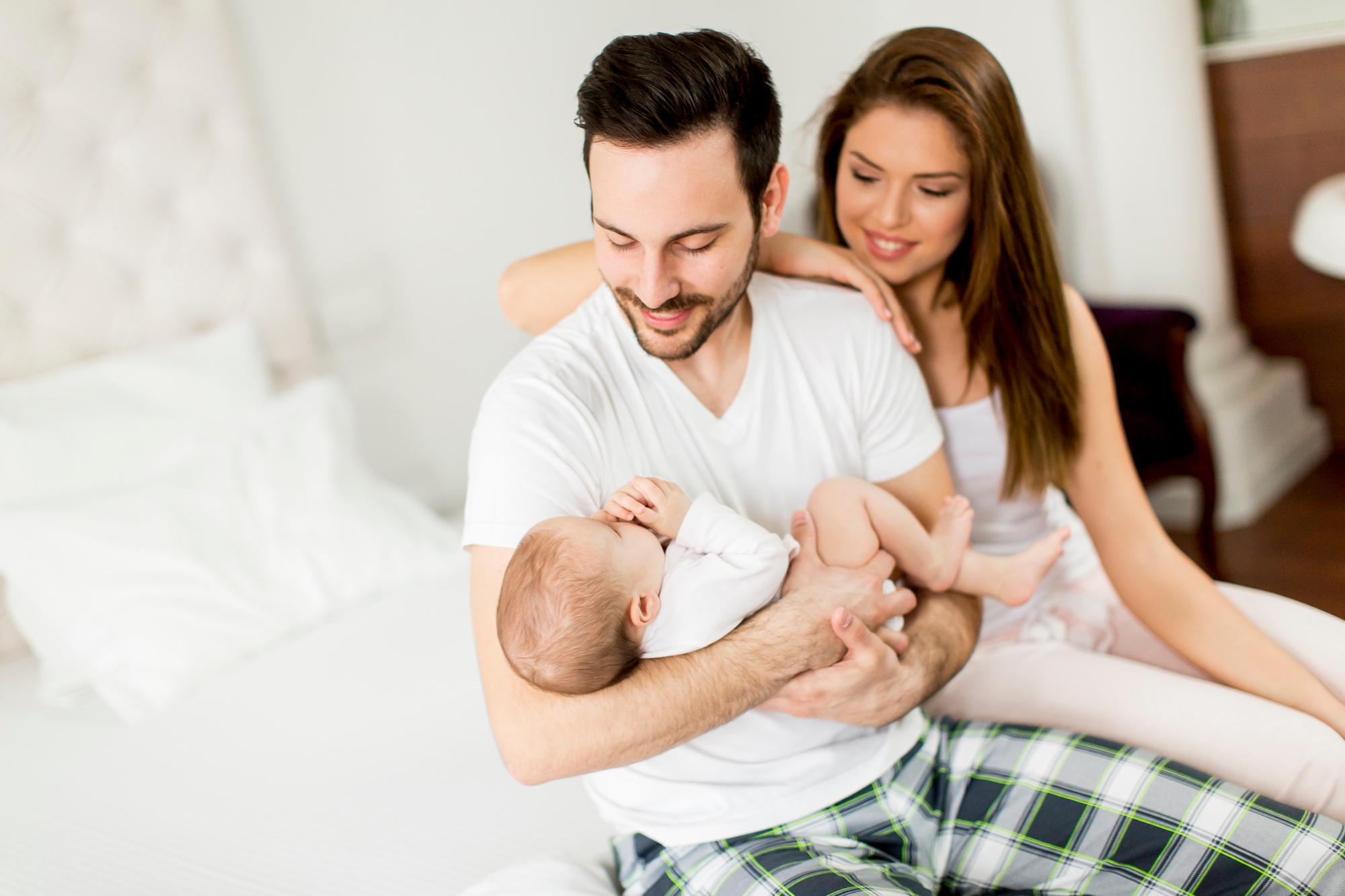 fisioterapia pediatrica Una pareja está sentada en una cama blanca y sonríe a un bebé que sostiene el hombre. Lleva una camiseta blanca y pantalones a cuadros. La mujer apoya la mano sobre su hombro. El entorno es un dormitorio luminoso y espacioso. Daganzo Adyre