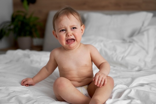fisioterapia pediatrica Un bebé sentado en una cama blanca, con aspecto molesto y llorando con la boca abierta y los ojos llorosos. La habitación tiene un fondo borroso con una planta verde y una cabecera de color marrón claro. El bebé lleva un pañal. Daganzo Adyre