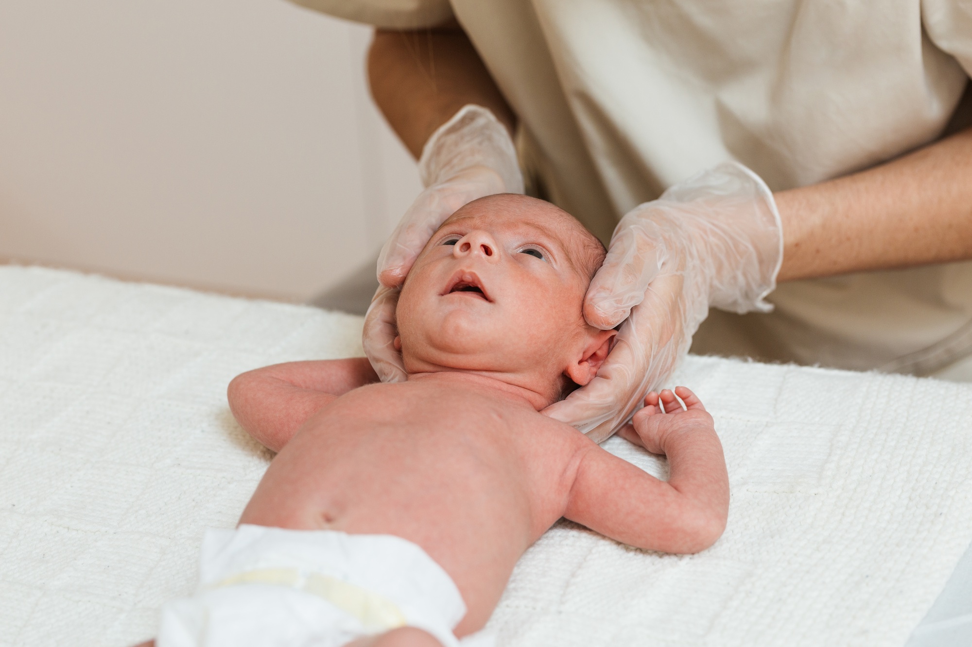 fisioterapia pediatrica Un bebé recién nacido está acostado sobre una superficie blanca mientras una persona con guantes sostiene suavemente la cabeza del bebé con ambas manos. El bebé lleva un pañal y mira hacia arriba. Daganzo Adyre