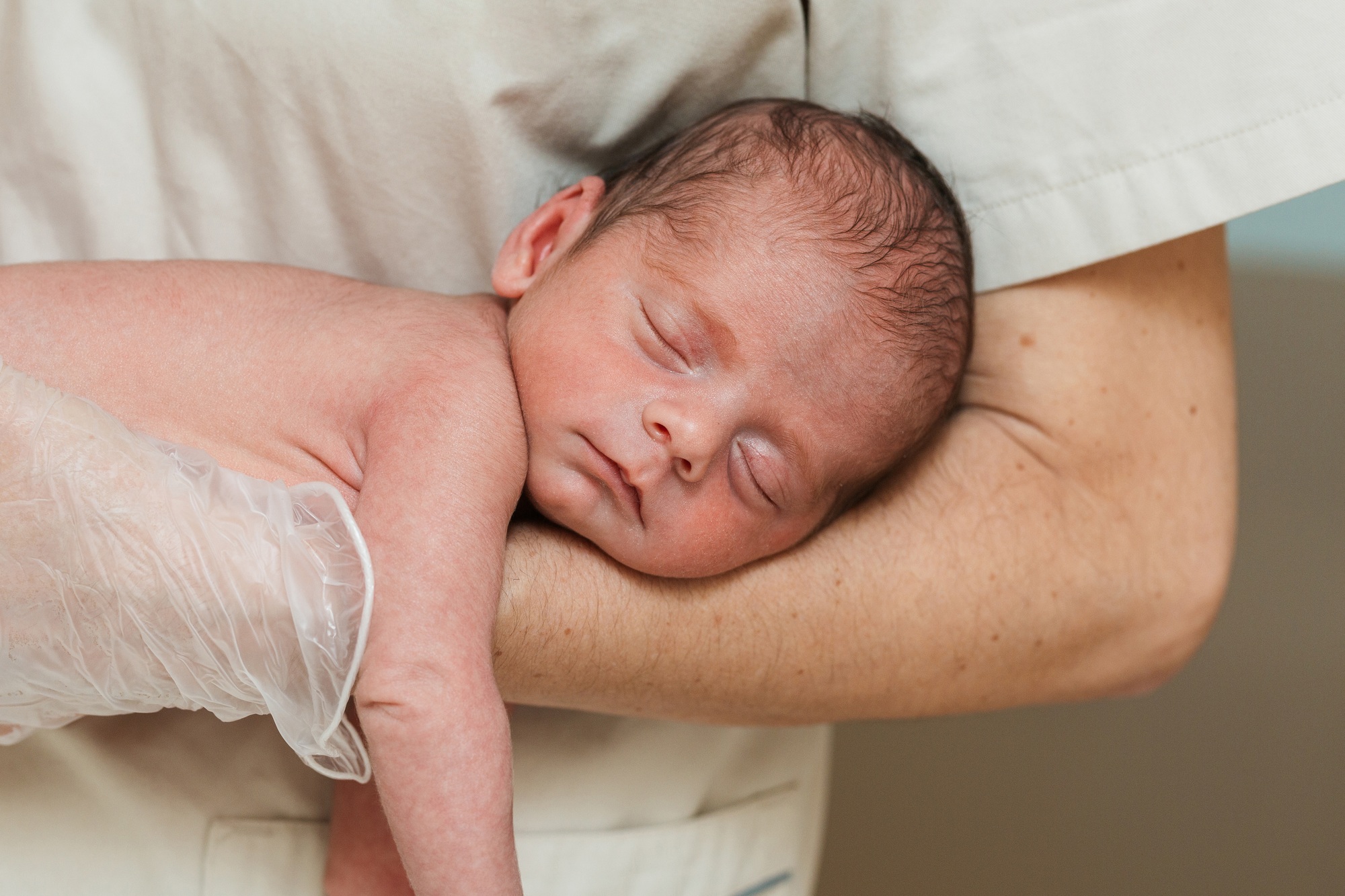 fisioterapia pediatrica Un bebé recién nacido duerme plácidamente en brazos de una persona que viste una camisa blanca. La persona sostiene al bebé con suavidad, con una mano que lleva un guante transparente. El bebé tiene un pelo fino y está envuelto en un cómodo abrazo. Daganzo Adyre