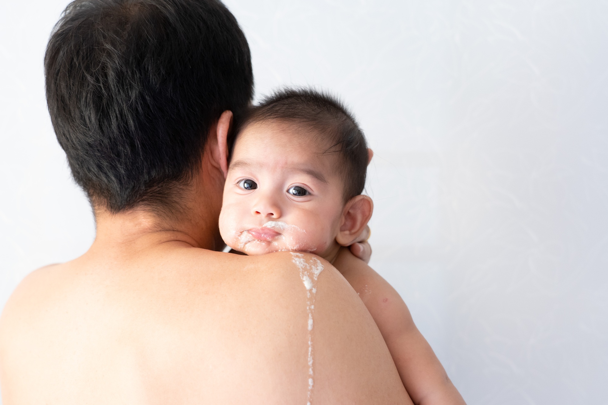 fisioterapia pediatrica Un bebé descansa sobre el hombro de un adulto y lo mira con los ojos muy abiertos. El bebé tiene restos de leche en la cara y el adulto tiene el pelo corto y oscuro. El fondo está ligeramente desenfocado. Daganzo Adyre