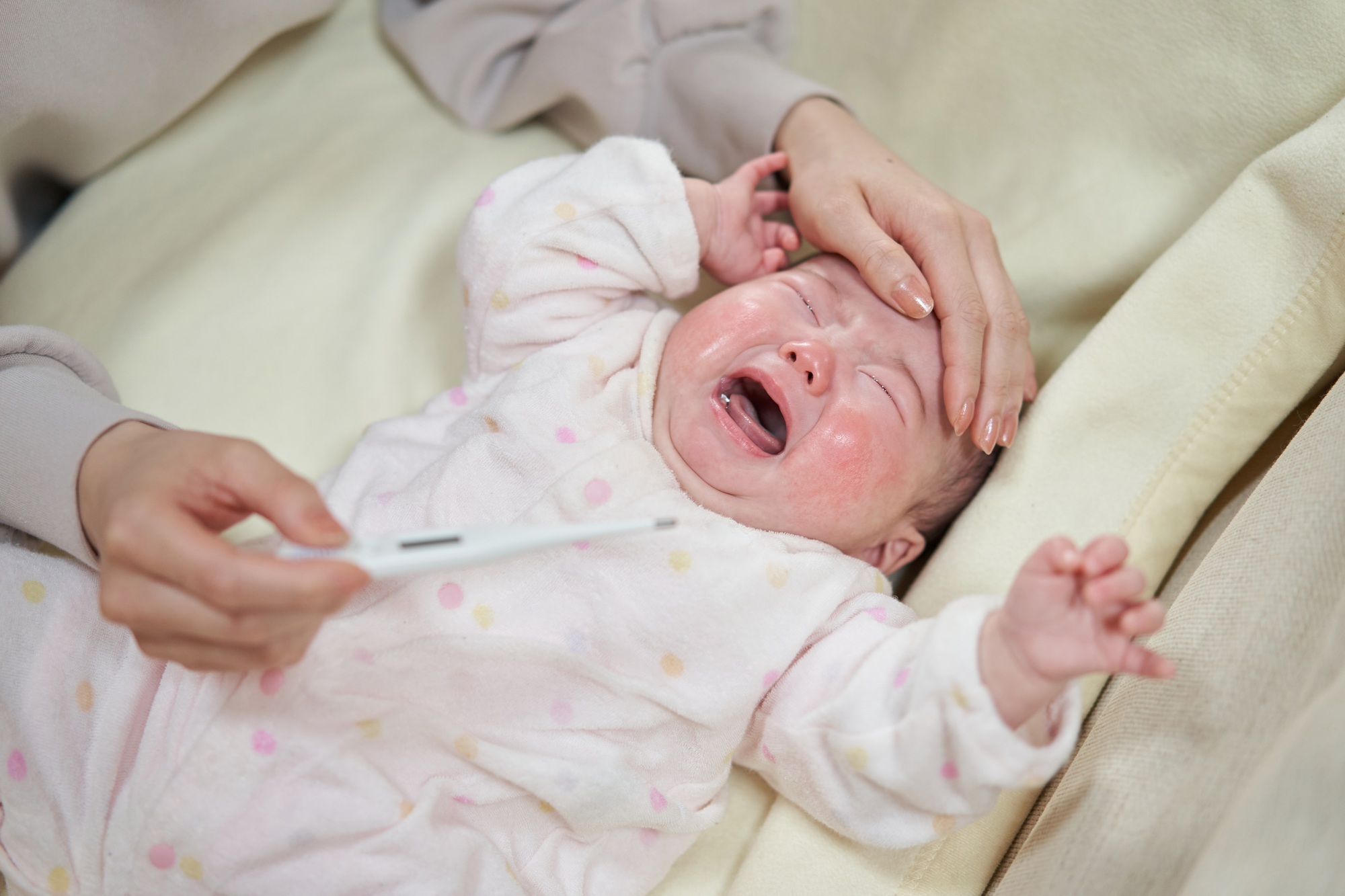 fisioterapia pediatrica Un bebé llora acostado en una cama, vestido con un mono blanco con lunares pastel. Las manos de un adulto tocan suavemente la frente del bebé y sostienen un termómetro, lo que sugiere que están controlando su temperatura. Daganzo Adyre