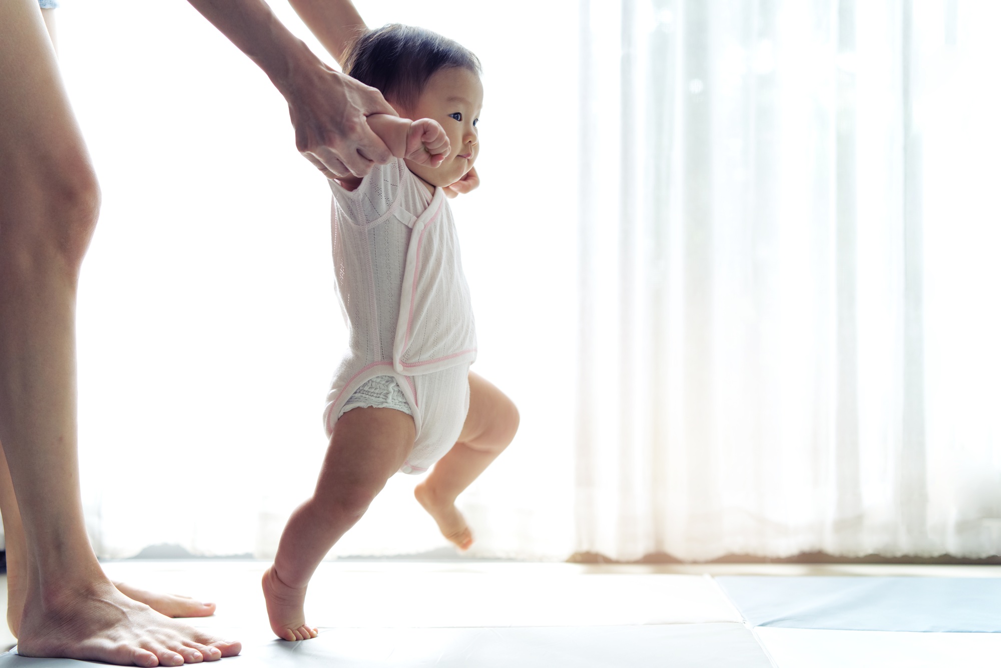 fisioterapia pediatrica Un niño pequeño con un traje blanco da pasos con la ayuda de las manos de un adulto sobre un fondo claro con cortinas transparentes. Se ven las piernas del adulto y los pies del niño sobre un suelo de color claro. Daganzo Adyre