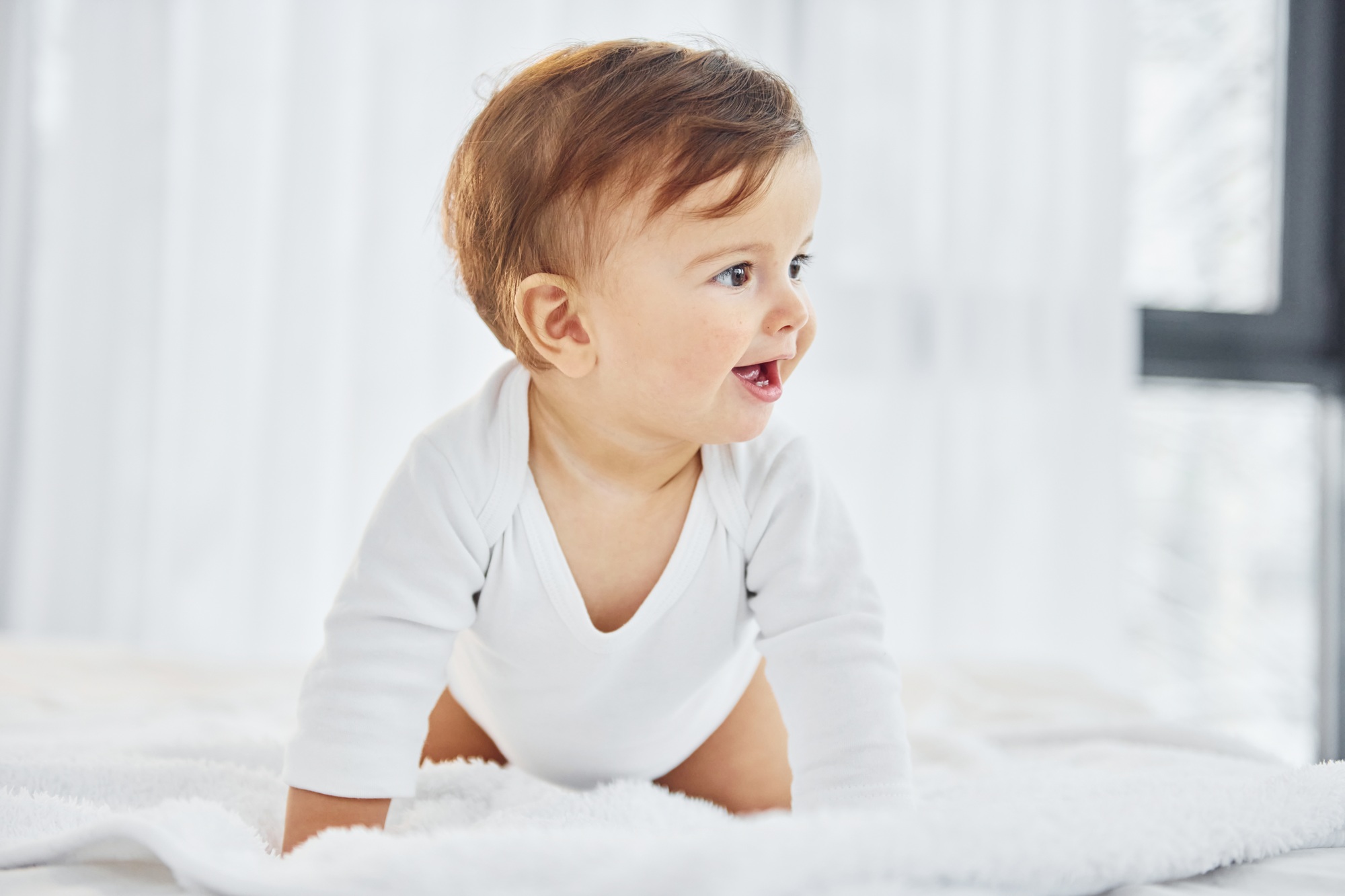 fisioterapia pediatrica Un bebé con un mono blanco sonríe y gatea sobre una manta blanca y suave. El fondo está suavemente iluminado con cortinas transparentes, lo que le da una sensación de luminosidad y amplitud. Daganzo Adyre